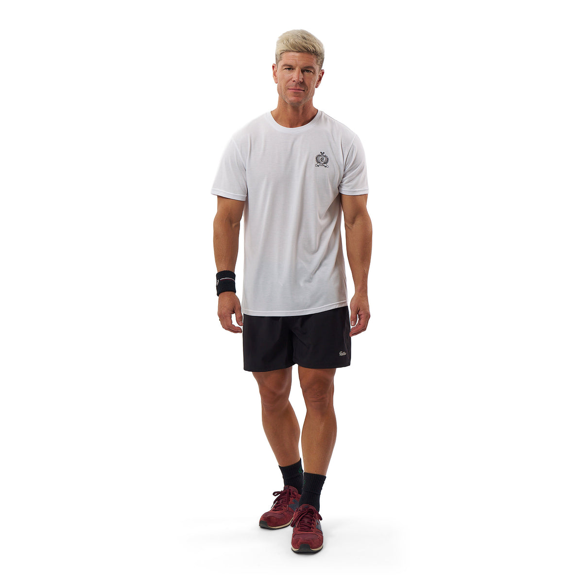 A man modeling a white short-sleeve athletic t-shirt with a small black chest logo, paired with black athletic shorts, black socks, and burgundy athletic shoes, against a solid white background.