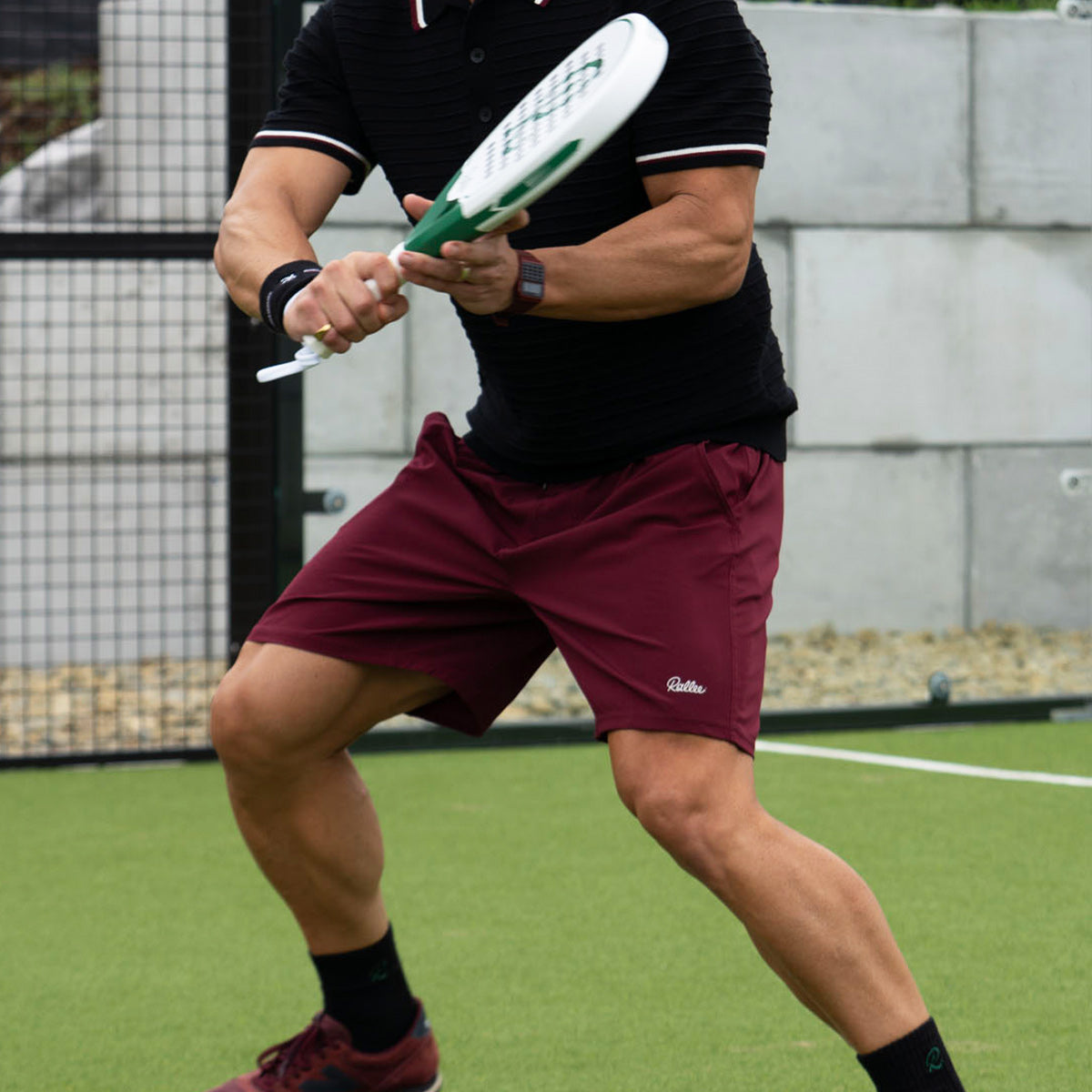 A man on a tennis court wearing burgundy athletic shorts, a black polo shirt, and black socks, holding a tennis racket in a ready stance.