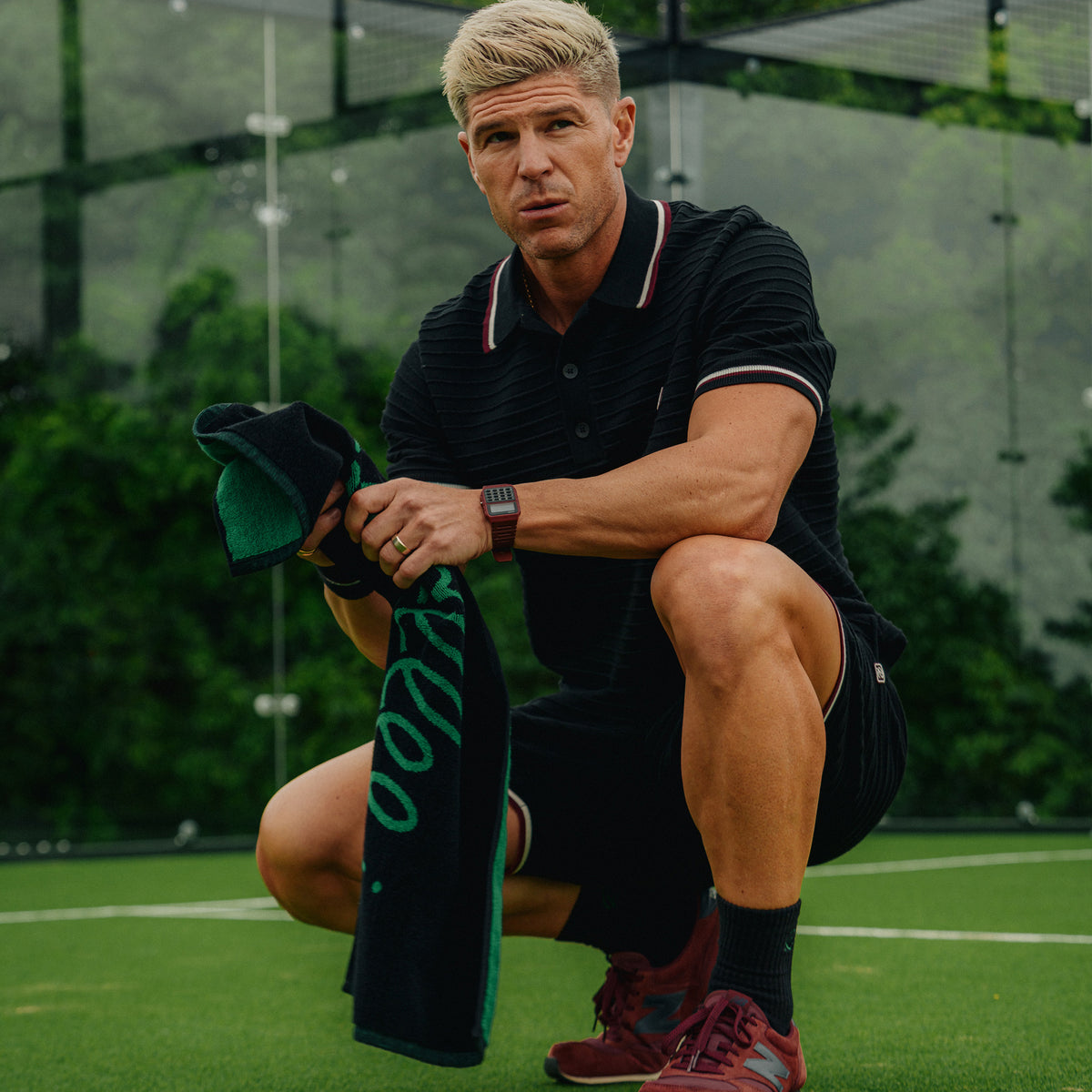 A man crouching on a tennis court holding a black and green rallee towel, wearing a black polo shirt with white and red trim, black shorts, and red sneakers.