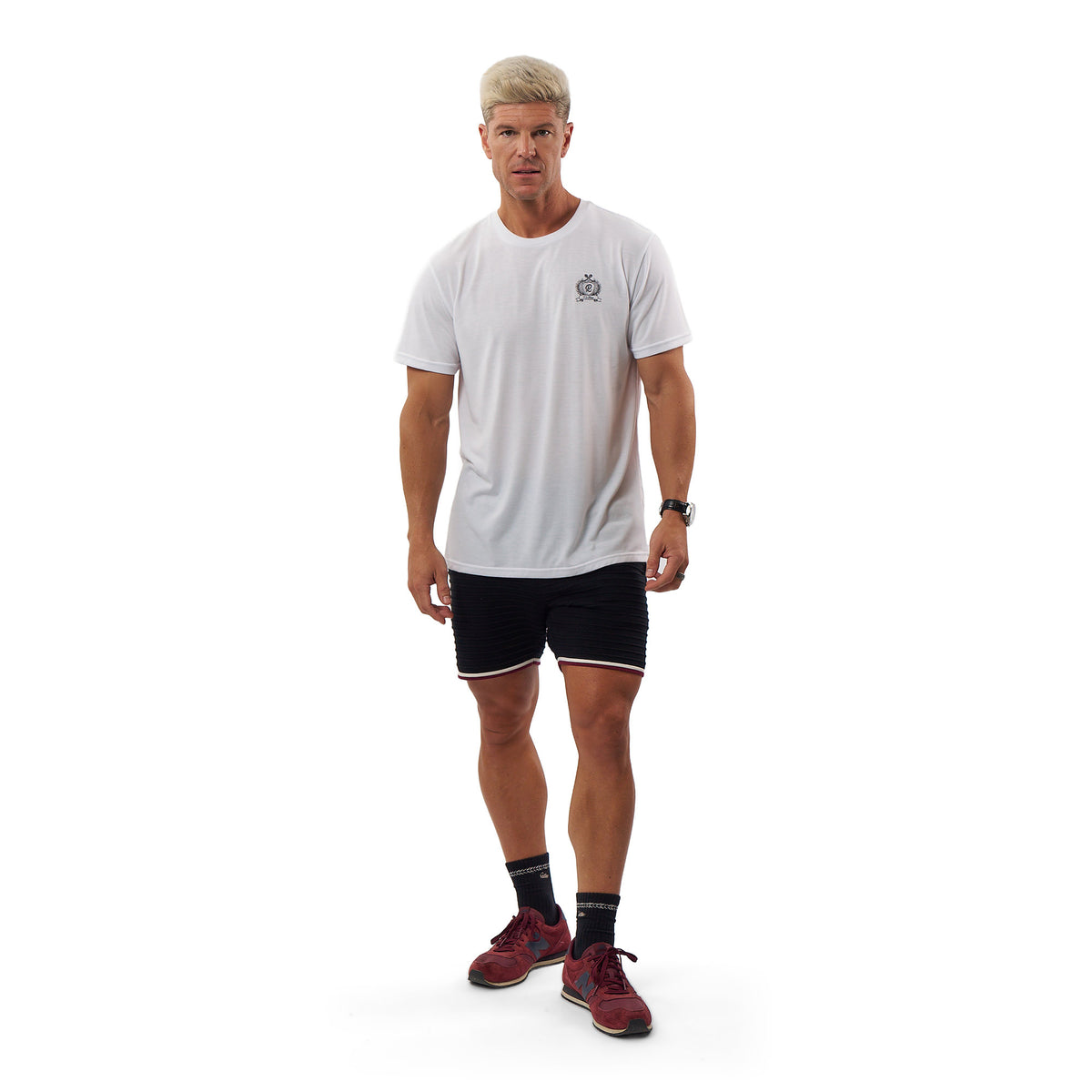 A man modeling a white short-sleeve athletic t-shirt with a small gray chest logo, paired with black athletic shorts, black socks, and burgundy athletic shoes, against a solid white background.