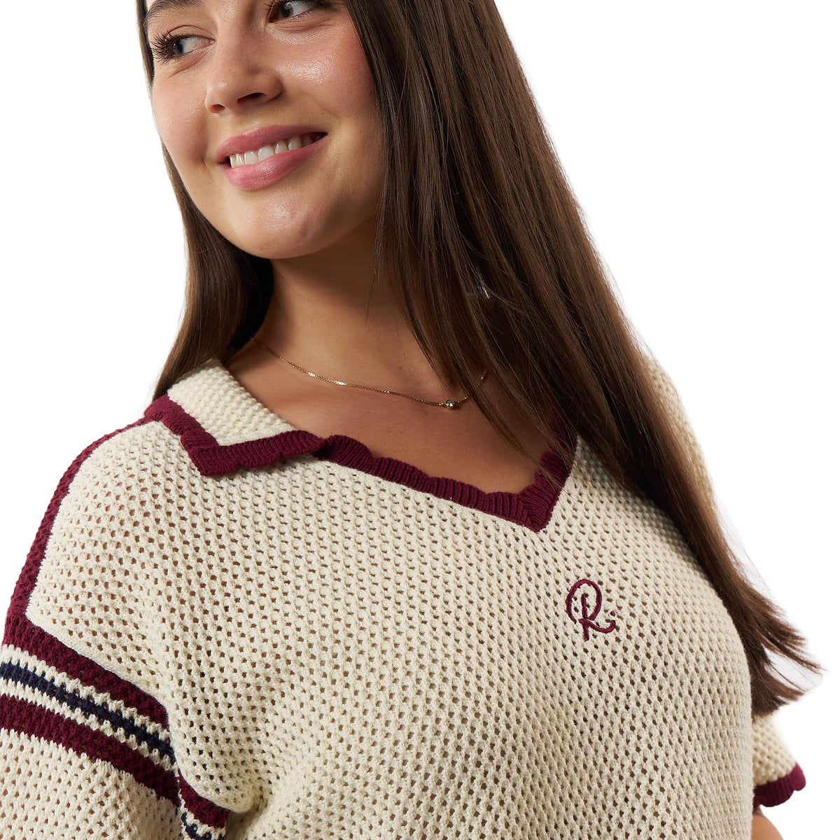 A close-up of a woman wearing a cream-colored knit sweater with burgundy trim and a small embroidered logo on the chest, set against a plain white background.