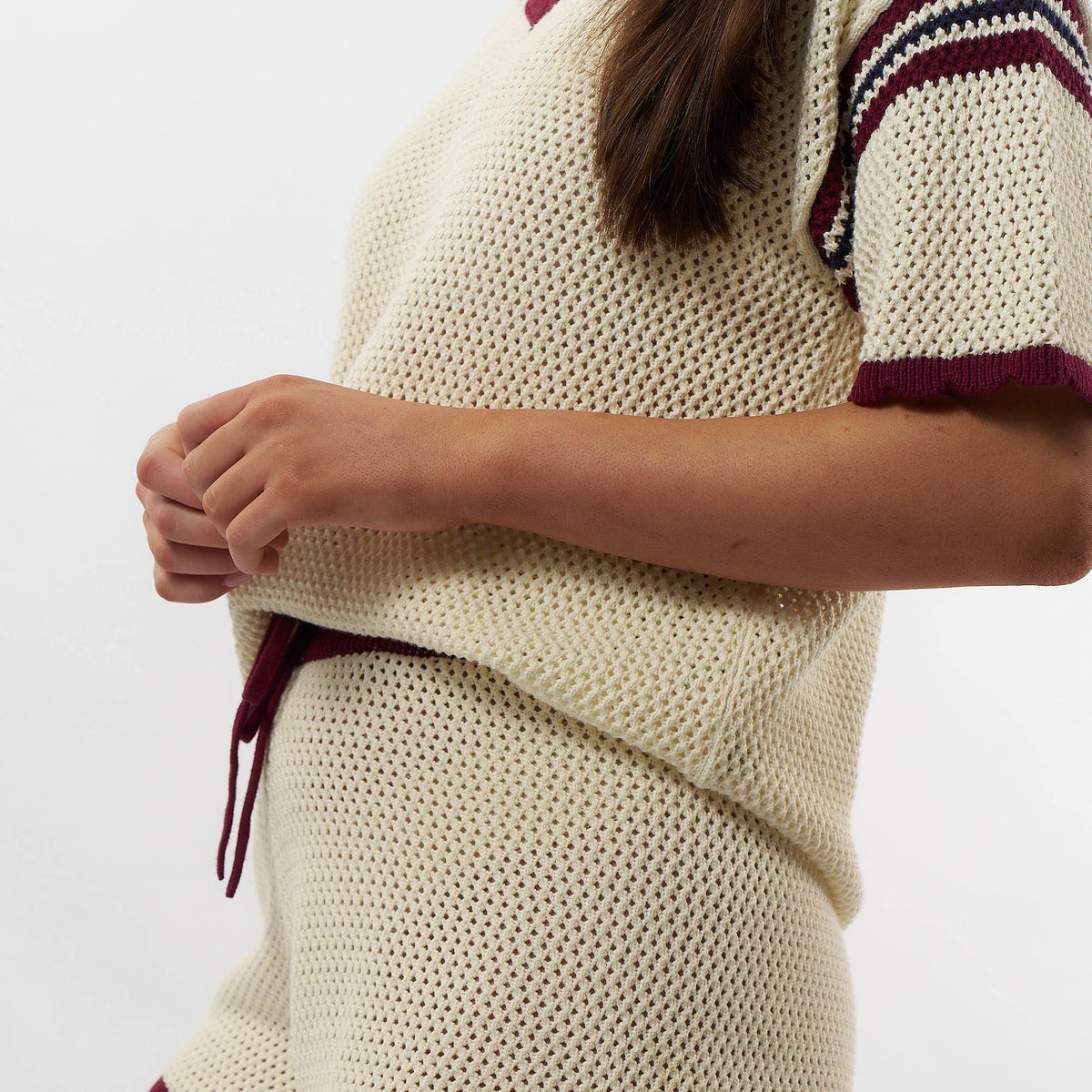 Close-up of a person wearing a chunky knit cream sweater with burgundy trim and a drawstring, against a plain light background.