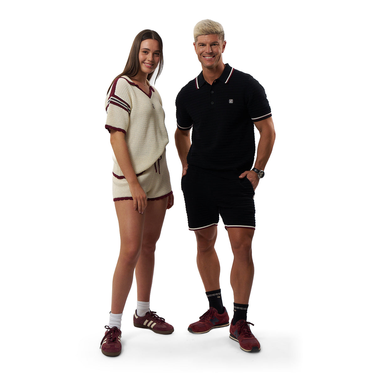 A man and a woman modeling retro-style athletic clothing against a plain white background. The woman wears a cream waffle-knit short-sleeve top and shorts with maroon trim, and the man wears a black short-sleeve polo shirt and shorts with white trim.