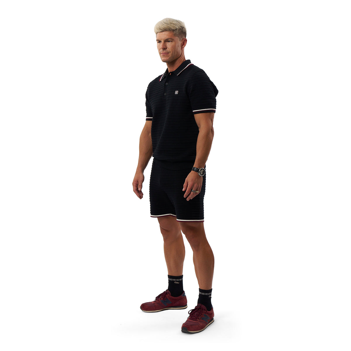 A man modeling a black athletic polo shirt and matching shorts with white trim, paired with dark socks and red sneakers, on a plain white background.