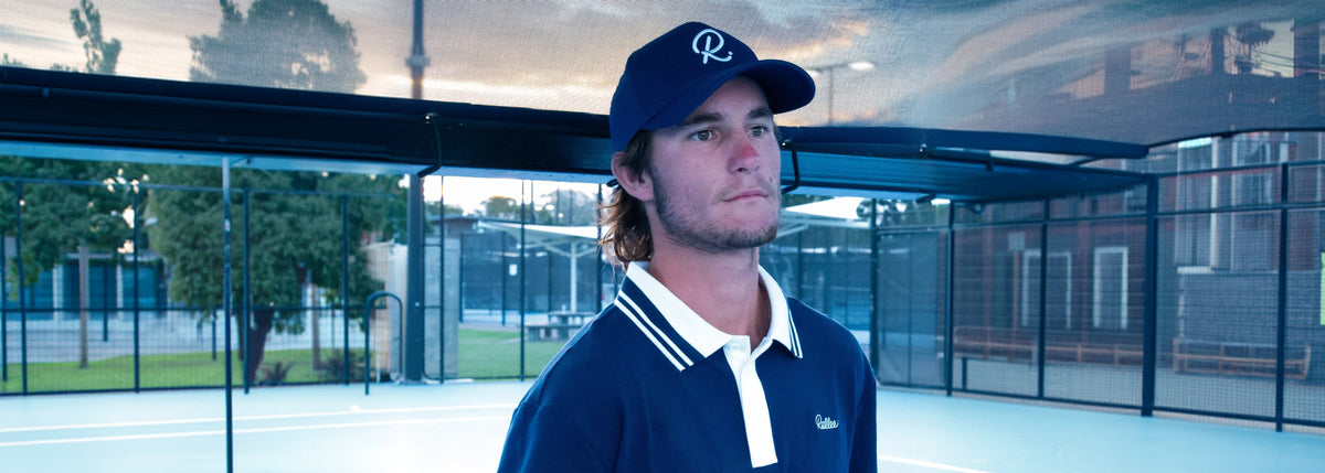 A man wearing a navy blue Rallee polo shirt and a matching navy cap with a white logo, standing on an outdoor tennis court.