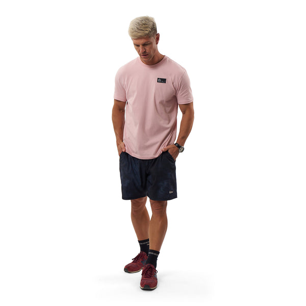 A man modeling a light pink crewneck t-shirt with a small black chest logo, paired with dark navy athletic shorts, dark socks, and burgundy athletic shoes, against a solid white background.