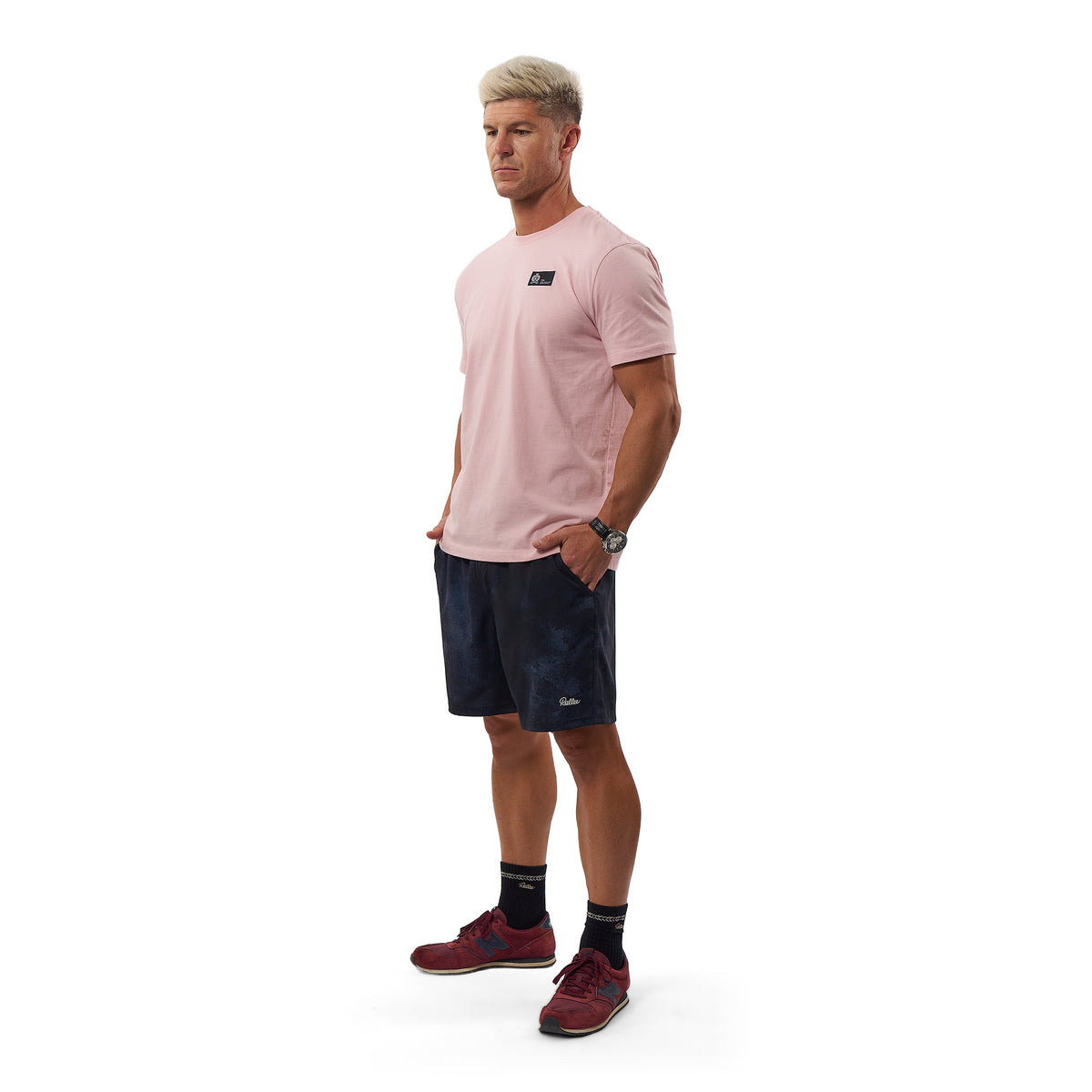 A man wearing a light pink athletic t-shirt, dark navy patterned shorts, black socks, and red running shoes, standing against a solid white background.