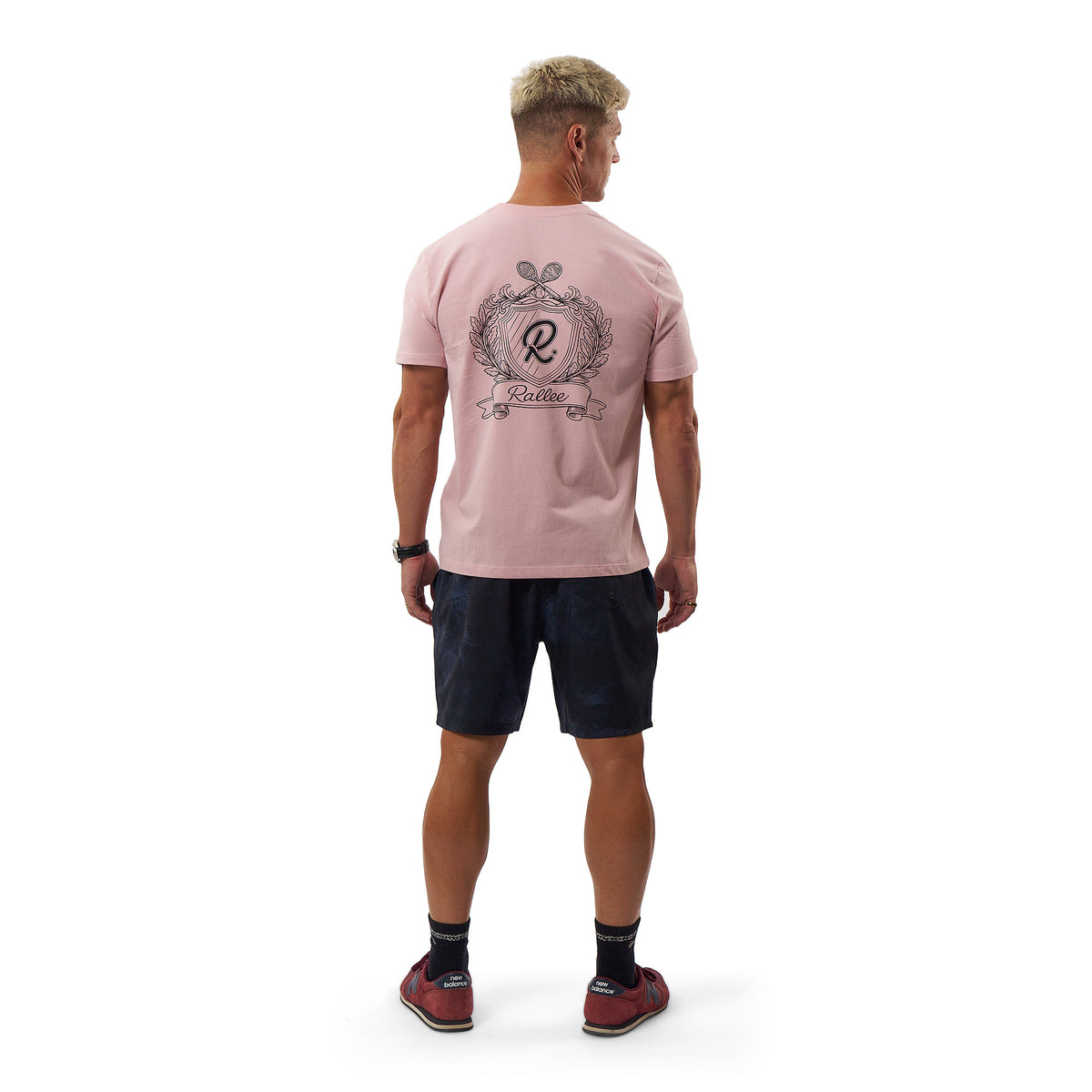A man viewed from behind wearing a light pink t-shirt with a large graphic print on the back, paired with dark shorts, socks, and red shoes, against a plain white background.